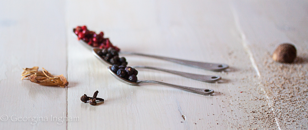 Whole peppercorns and pink peppercorns measured in metal spoons with cloves, nutmeg and ground spices scattered on a white table — classic aromatics used in cooking with herbs and spices.